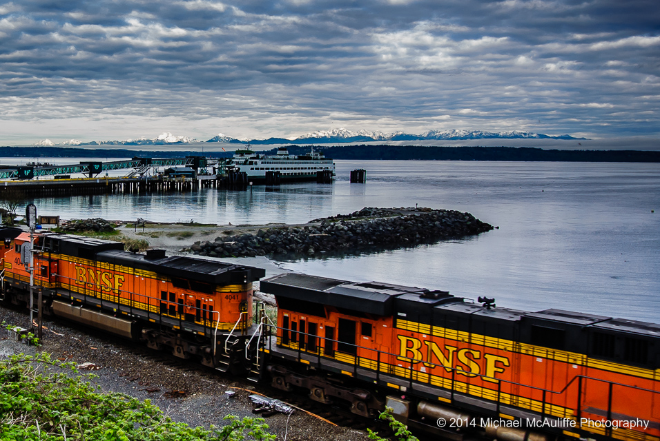 The sun coming up on the Olympic Mountains from Edmonds, WA  with a ferry and a train in the foreground.